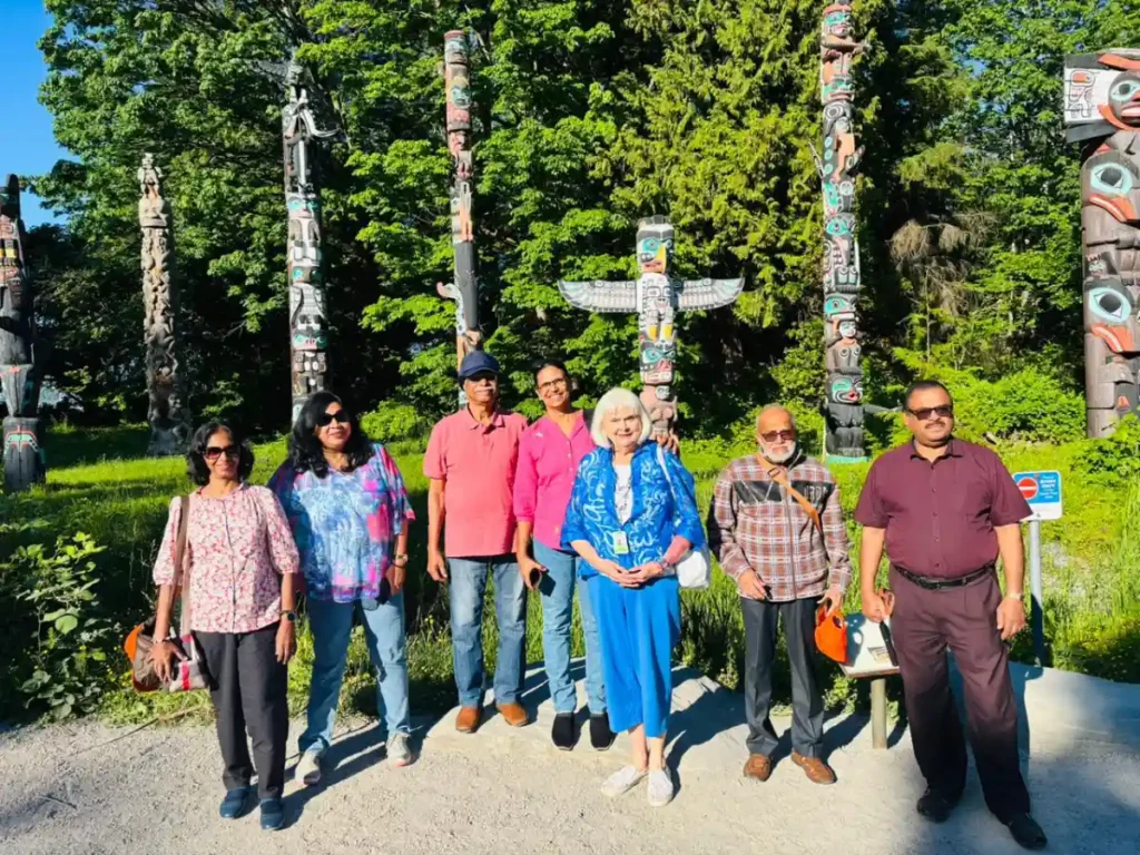 Visitors posing at the Stanley Park totem poles in Vancouver, Canada with carved wooden poles behind them.